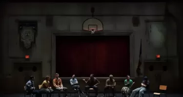 A group of people sit on wooden stacking chairs in a semi circle on a dark stage made to look like a community auditorium. Behind them is a run down wall, stage with red curtains, basketball hoop and a banner.