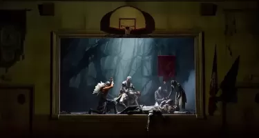 A group of people form a tableau pose on a stage that looks like it is in a community auditorium. They are dressed in medieval armour and costume. One is lying down as if dead. A basketball net hangs above the stage.