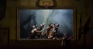 A group of people form a tableau pose on a stage that looks like it is in a community auditorium. They are dressed in medieval armour and costume. One holds a knife. A basketball net hangs above the stage.