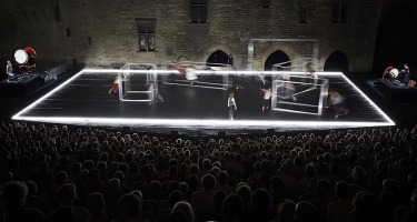 A person stands on stage with their back to the audience watching pairs of people push around five hollow metal boxes. The boxes are blurred with the motion.