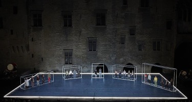 Groups of people standing in five hollow metal boxes arranged on stage in a semi circle facing outwards.