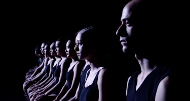 Batsheva Dance Company. Eleven fair skinned dancers in tight black sleeveless tops and shorts sit side by side in a line with their hands on their knees. They are facing to the left. The dancer on the far right is closest to the camera with the others receding back into the frame to the left.
