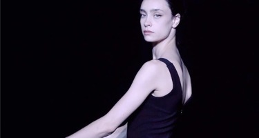 Batsheva Dance Company. A portrait from the waist up of a fair skinned woman with black hair tied back. Her body faces left with hands slightly outstretched, and her head is turned to face the camera. She is wearing a tight, sleeveless black top and standing against a black background.