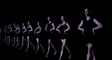 Batsheva Dance Company. Eleven fair skinned dancers in tight black sleeveless tops and shorts stand side by side in a line with their hands on their hips. They are facing slightly to the left of the frame. The dancer on the far right is closest to the camera with the others receding back into the frame to the left.