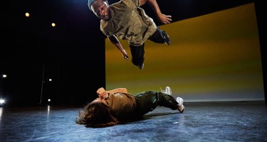 A Black male dancer in a baggy olive t-shirt and blue toque is suspended horizontally in the air overtop of a person with long brown hair lying on the ground.