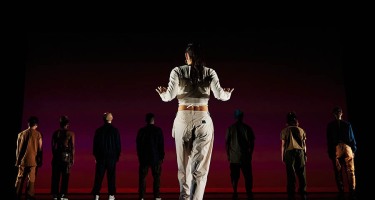 Light skinned woman with long dark hair, dressed in a long sleeve white crop top, baggy white pants and sneakers stands at the front of a stage with her back to the camera and arms slightly raised. A row of seven people in shadow stand at the back of the stage.
