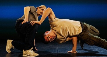 A light skinned male dancer with a dark moustache, beige cap, olive green t-shirt, dark pants and light coloured running shoes crouches backwards on stage. Another light skinned male dancer with dark hair, beige t-shirt, and olive green pants supports himself with one hand on stage, one hand on the other man's knee and outsretched foot.