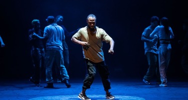 A black, male dancer in a beige t shirt, black cargo pants and sneakers dances centre stage with arms raised slightly in front of him. Six other people linger in the in the backgound behind him.