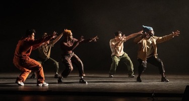 Five dancers in neutral coloured baggy clothing stand in an offset row, slightly crouched with their arms outstretched.