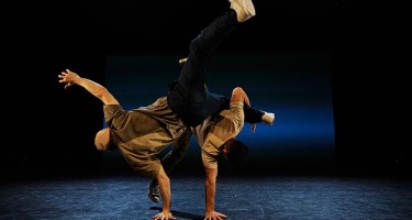 Two male dancers each balance on one hand, with their legs raised in the air. One man clasps the other's left leg with the crook of his arm.