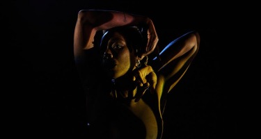 A portrait photo of a Black woman, largely in shadow with highlights striking her from the side. She is wearing a black sleeveless top and is posing with one arm draped over her head with the other bent upwards beside her face.