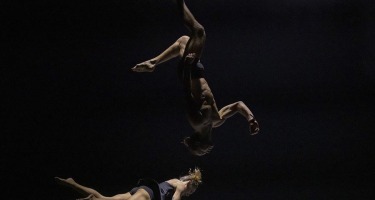 Dancer doing aerial backflip over another dancer on ground.