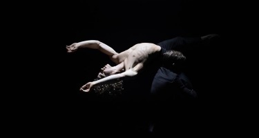 Male dancer being held up by a person in shadow. He is bent over backwards with his arms stretched out behind him.