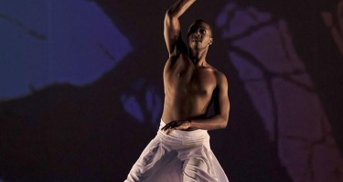 A lone Black, male dancer dressed in flowing white pants faces the camera with one arm at his hip and one raised above his head. The backdrop is dark blue with the silouetted outline of a leafless tree.