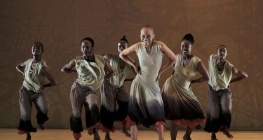 Six Black dancers, two women and four men, stand in a group facing the camera with hands skewed on hips.. They are all wearing sleeveless tops and flowing pants or skirts in various neutral colour tones.  The backdrop is medium brown.