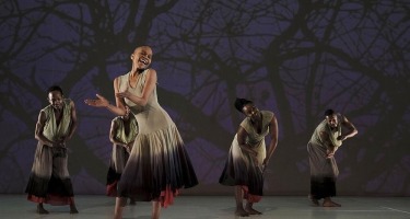 Five Black dancers - two women and three men - stand facing the camera  They are all wearing sleeveless tops and flowing pants or skirts in various neutral colour tones. The woman in the front has her hands raised as if about to clap. A silouette of tree branches with no leaves is projected on the wall behind them.