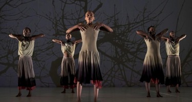 Five Black dancers - two women and three men - stand facing the camera with their arms raised in front of their chests. They are all wearing sleeveless tops and flowing pants or skirts in various neutral colour tones. A silouette of tree branches with no leaves is projected on the wall behind them.