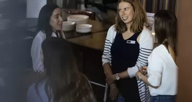 DanceHouse One social event. A group of four women with long brown hair stand in a circle chatting. A table with stacks of plates and a stand up menu is in the background.