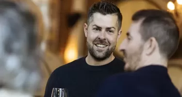 DanceHouse One social event. Two light skinned men with short brown hair and black tops chat with each other. One has a light beard and is holding a glass of white wine.