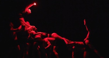 A group of people form a tableau that looks like a wave made up of multiple bodies. The person at the peak arches backwards and hols a light. Everyone is lit in red against a black background.
