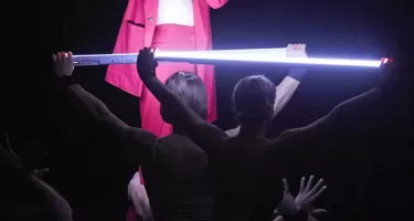 A woman in red pants, and a red blazer open down the front to reveal a patterned sports bra speaks into a wireless handheld microphone. She is being raised up by another dancer and has one arm raised above her head. Two dancers hold two flourescent tubes horizontally above their heads.