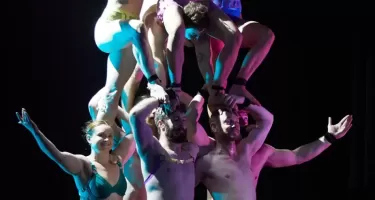 A group of dancers in brightly coloured underwear form a human pillar, with one group standing on the shoulders and heads of the other.