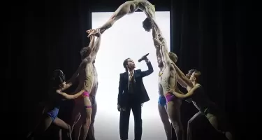 A woman in an oversized dark suit, white shirt and tie speaks into a handheld wireless microphone. Another group of dancers in brightly coloured underwear form a human archway, framing the woman in a suit. A bright, white, door-like rectangle is in the background.