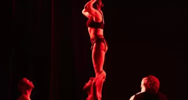 Profile photos of a woman standing on a man's shoulders, while another woman does an arc-ing handstand on the middle woman's shoulders, creating a pillar of three people. Three others stand below looking up. They are all lit with red light against a black background.