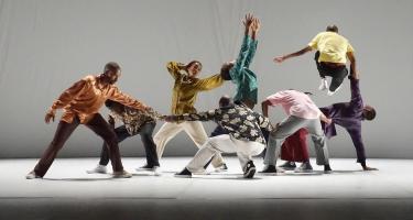 Group of colourfully dressed dancers with some crouching and reaching for each other, some reaching upwards and one jumping.