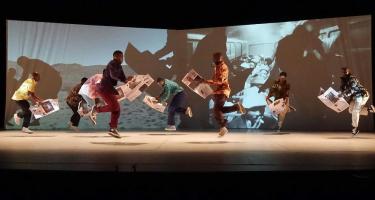 Via Katlehong Dance Company. Eight colourfully dressed dancers spread across the stage in various states of jumping. All are holding broadsheet newspapers open in front of them. Black and white images of a desert landscape paired with people riding on a train are projected behind.