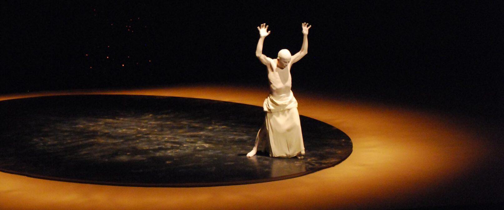 Sankai Juku. Figure covered in white body paint with a white wrap skirt stands in a dark circle, with arms raised.