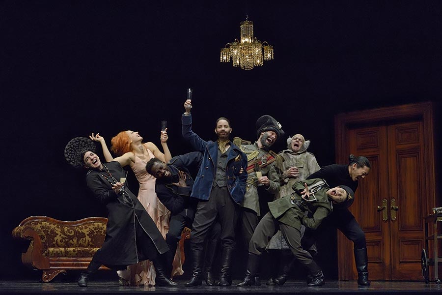 Group of dancers in military and period Russian costumes on stage with chandelier.