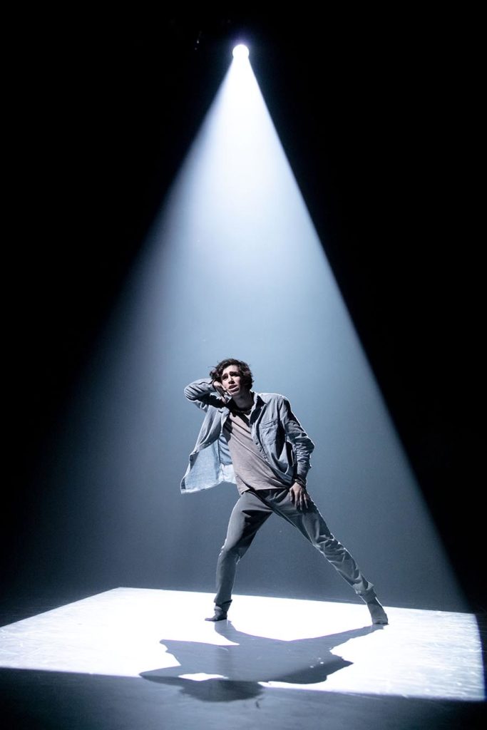 The Friday Round-Up 1 Male dancer in spotlight with hand raised to ear as if listening.