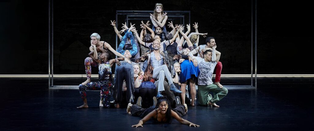 Babel. Group of people with arms raised closely surround man seated in chair.