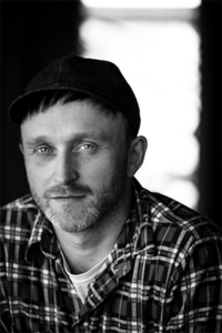 Portrait of a man facing forward wearing a white t-shirt and plaid button up top.