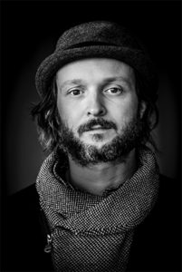 Portrait of bearded man facing forward wearing a Trilby had and a patterned scarf.