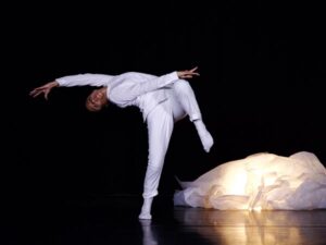 Zahra Shahab, thaw, photo by Yvonne Chew. Dancer dressed in white pant suit on one leg arching backwards. A crumpled pile of cloth sits in the bottom corner on the floor.