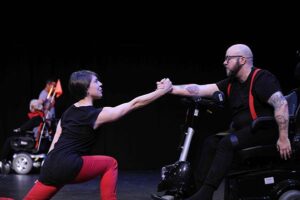 All Bodies Dance Project. Woman in black short sleeve shirt and red pants on one knee reaches out and clasps a hand with a man with a crutch wearing black clothes with red suspenders.