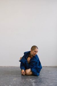 Shion Skye Carter, photo by Lula-Belle Jedynak. Woman with shaved head crouches on ground with arm wrapped around her knee.