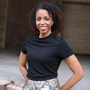 Marua Garcia. Smiling woman with dark, curly, shoulder length hair in a black t-shirt and floral shorts leans against a waist high brick wall with her hands behind her back.