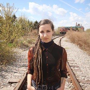 Sophie Dow. Woman with sandy brown braid and three quarter length brown blouse stands on railway tracks facing forward with her hands in her pockets.