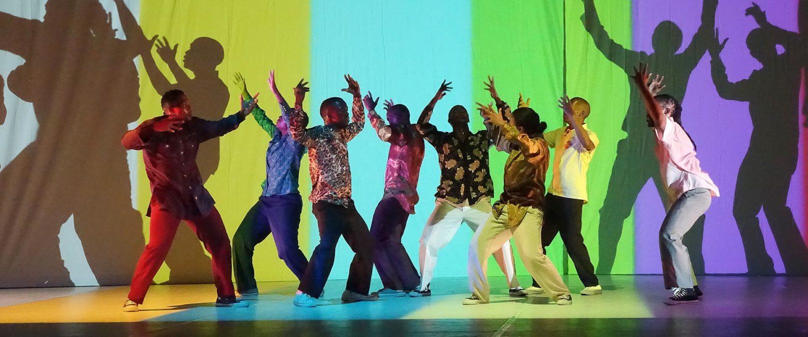 Via Katlehong Dance Company. Eight colourfully dressed dancers stand closely in a line with their arms raised. Behind them is a bright projected backdrop of wide bands of colour.