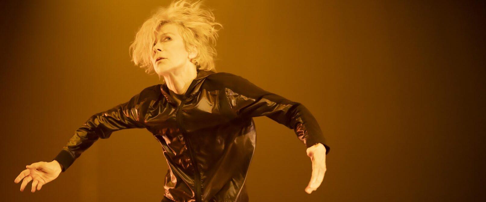 A woman with shaggy blond hair and a shiny black jacket faces the camera with her arms outstretched to her sides.