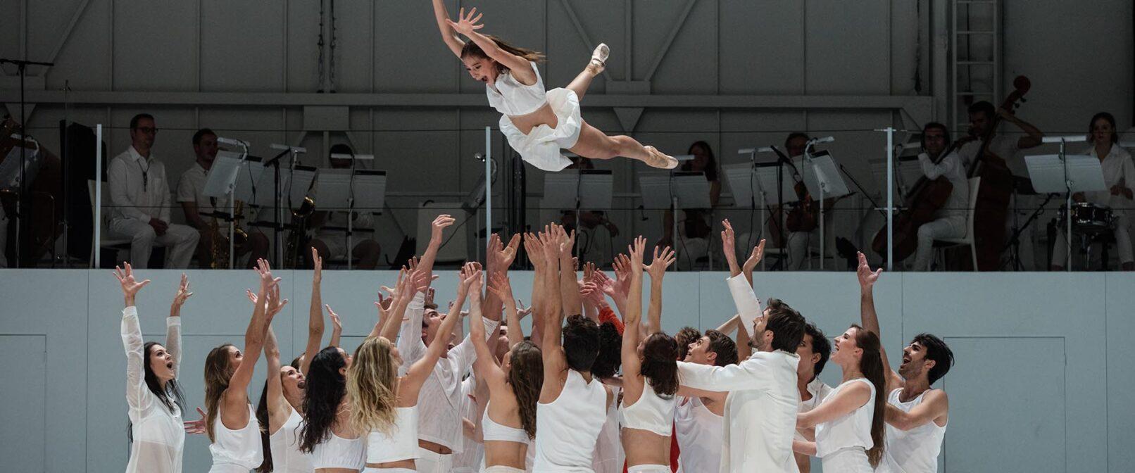 Paris Opera Ballet - Play. Large group of dancers dressed in white clothing stand in a group, a female dancer in white is thrown in the air by the group.