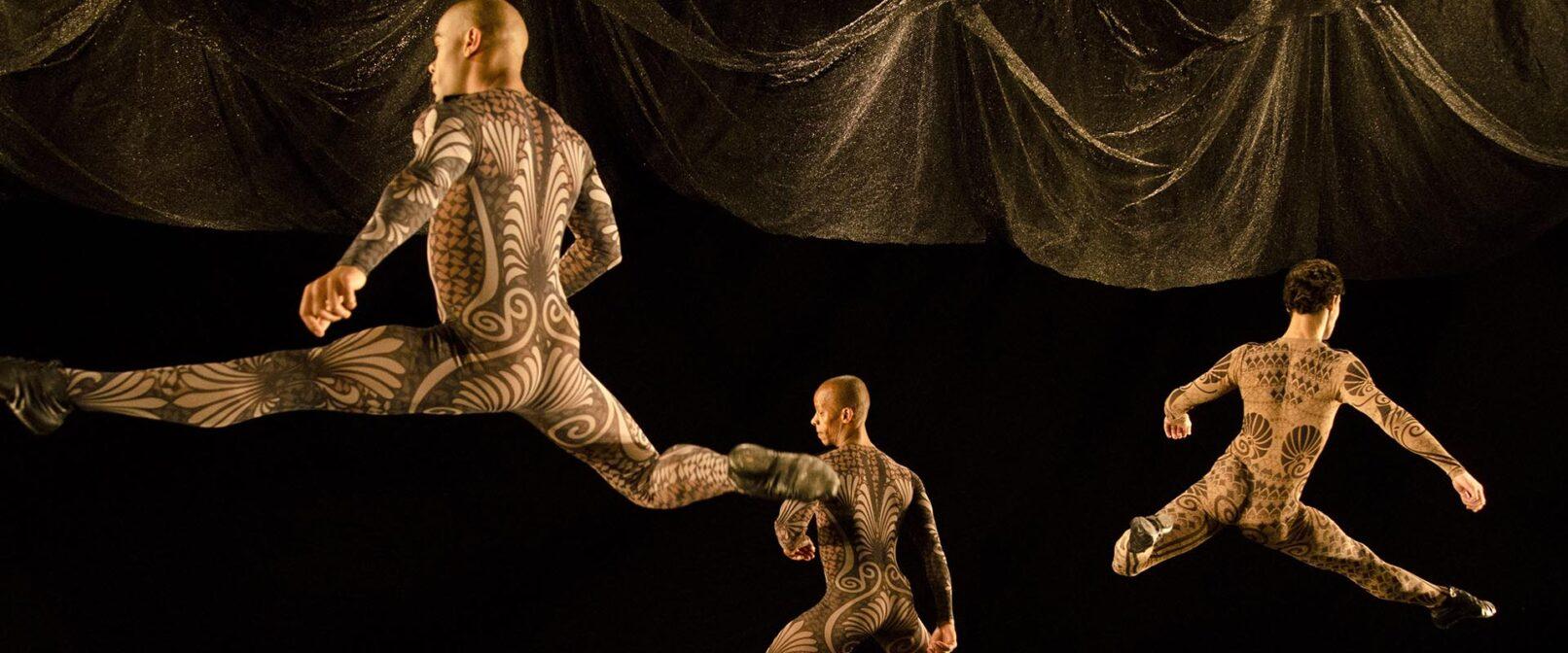 Grupo Corpo. Sem Mim. Three male dancers in skin tight leotards that resemble body tattoos leap on stage against a black backdrop and dark gold coloured curtain.