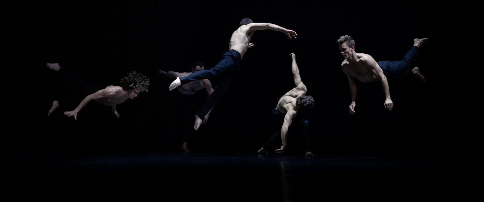 Circa. A group of five, shirtless, fair skinned male dancers in dark blue trousers. Three leap so that their bodies are parallel to the stage and the other two bend forward with arms stretched downward. They are lit against a dark background.