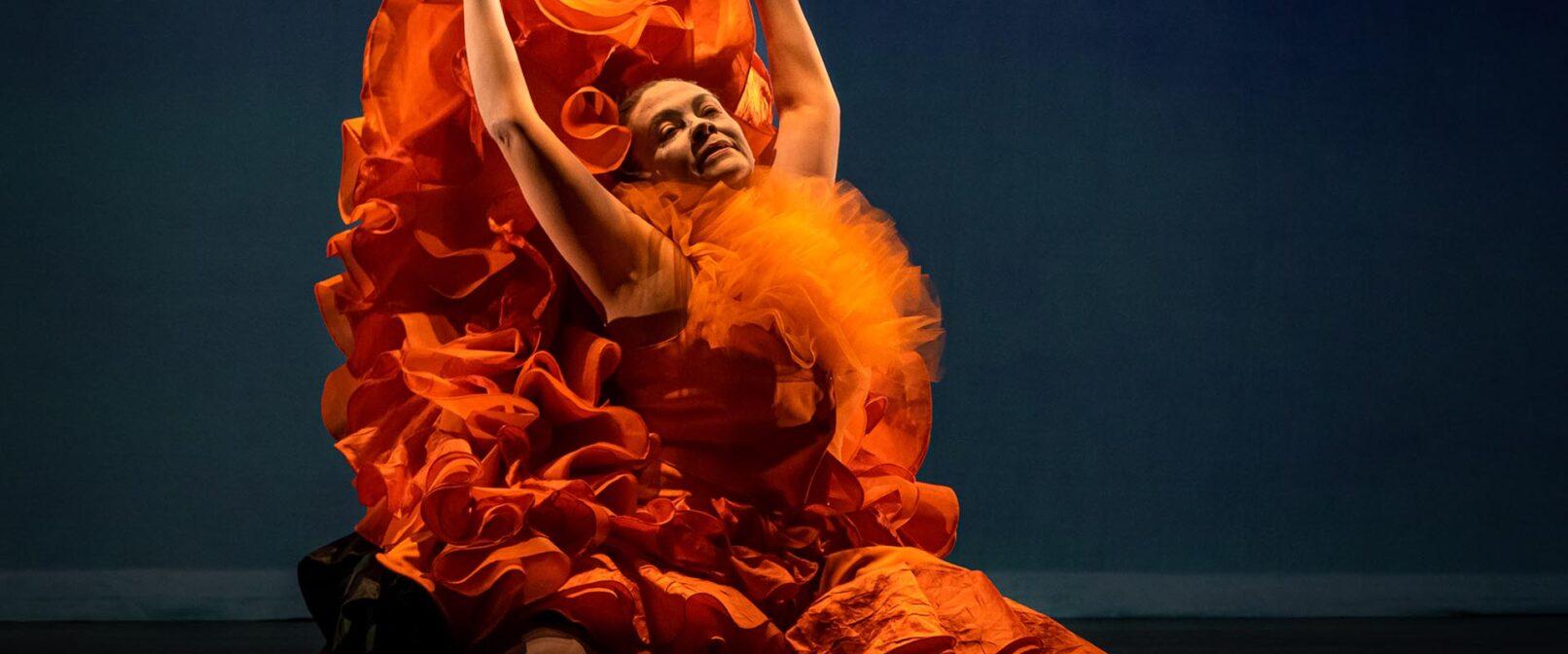 Bailando. Person in ruffled orange chiffon top with full, ruffled orange skirt kneels with arms raised high, holding edges of the skirt.