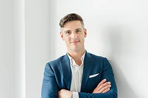 Kyle Bienvenu. Portrait photo of a light skinned man with short brown hair. He is wearing a blue blazer and white button-up shirt and has his arms crossed across his chest.