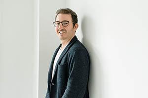 Chris Stoff. Portrait photo of a light skinned man with short brown hair and dark rimmed glasses. He is wearing a dark blue blazer and white t-shirt and is leaning against a white wall.