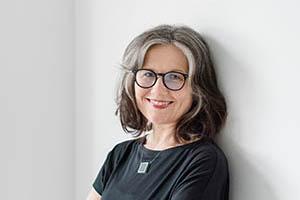 Liesl Jauk. Portrait photo of a light skinned woman with shoulder length, salth and pepper hair and black rimmed glasses. She is wearing a short sleeve black t-shirt and a necklace with a square shaped pendant.